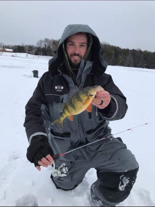 Fisherman in Striker Ice float suit holding a fish and fishing rod, kneeling on the ice